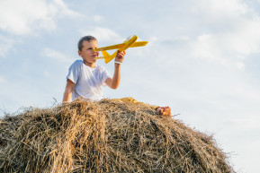 portrait-barefoot-boy-hat-haystack-field-playing-with-toy-airplane-light-sunny-day-outdoor